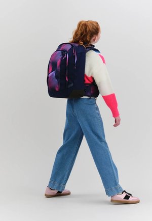 Young person with red hair wearing a colorful sweater, blue jeans, pink sneakers, and carrying a large dark backpack with purple and pink patterns.