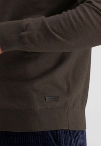 Donkerbruine langemouwen trui met geribde manchetten en zoom, met een klein zwart "pierre cardin" label nabij de taille boven een donkerblauwe corduroy broek.