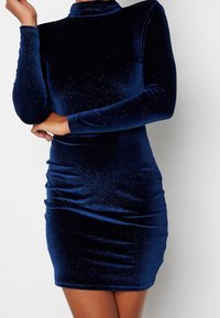 Femme portant une robe ajustée en velours bleu marine à manches longues avec des détails scintillants subtils sur un fond blanc uni.