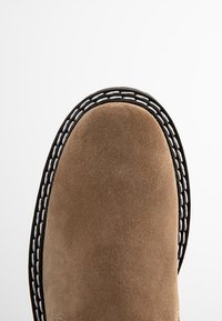 Bruine suede laars met een ronde neus, met zwarte randen en witte decoratieve stiksels langs de naad. Glad oppervlak.
