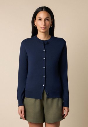 Donna con capelli lunghi e scuri che indossa orecchini a cerchio dorati, un cardigan blu navy con bottoni e pantaloncini verde oliva su uno sfondo beige.
