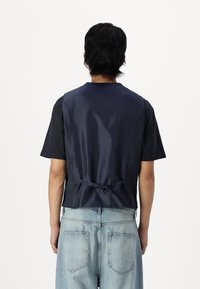 Mørkeblå ermeløs vest med glatt stoff bak, forsterkede sider og justerbar snøring i midjen. Brukt over en svart skjorte og jeans.