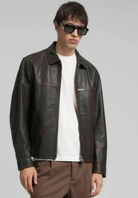 Giacca di pelle marrone con zip frontale, colletto appuntito e due tasche sul petto. Indossata sopra una maglietta bianca, abbinata a pantaloni marroni e occhiali da sole neri.