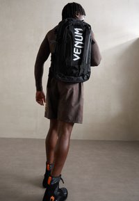Sac à dos noir avec logo "VENUM" blanc, plusieurs compartiments, bretelles rembourrées et surface texturée, porté par une personne en shorts de sport.