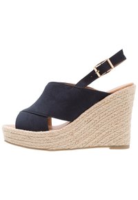 Marinblå kilklackssandal med öppen tå och urtag i ovandelen. Tillverkad av mjukt mockamaterial, med en vävd jute-liknande sula och justerbar ankelrem.