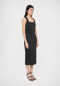 Čierne midi šaty s textúrovaným celoplošným monogramom MK. Majú štvorcový výstrih a sú skombinované s čiernymi sandálmi na opätku so remienkami.