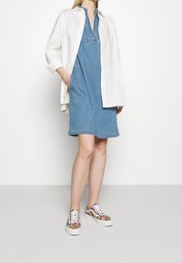 Abito di denim con colletto, maniche corte e tasca frontale, abbinato a scarpe da ginnastica colorate e a fantasia. Camicia bianca sovrapposta all'abito.