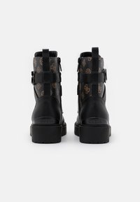 Un paio di stivali neri in pelle alla caviglia con suole spesse, parte superiore in tessuto stampato marrone, e "GUESS" impresso sul tallone posteriore.