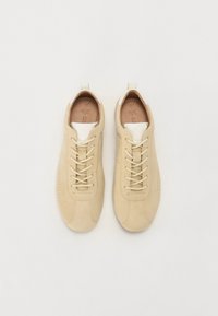 Beige semskinn-sneakers med en teksturert finish, hvit detalj på hælen og tradisjonell snøring. Sålen er flat, designet for hverdagsbruk.