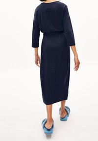 Granatowa, średniej długości sukienka o luźnym kroju, z rękawami do trzech czwarte oraz wiązanym paskiem w talii, w połączeniu z jasnoniebieskimi, puszystymi klapkami.