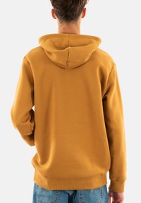 Hoodie giallo senape con cappuccio regolabile, polsini e vita a coste. Tessuto morbido e vestibilità rilassata, abbinato a jeans in denim blu.
