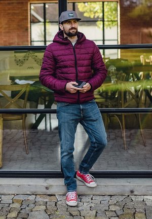 Man in maroon puffer jacket, gray cap, jeans, and red sneakers leaning against glass window, holding a wallet outdoors.
