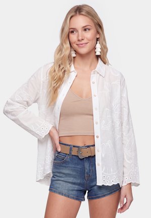 Młoda kobieta z długimi blond włosami, ubrana w białą haftowaną koszulę zapinaną na guziki, na beżowym crop topie oraz niebieskie dżinsowe szorty z brązowym paskiem.