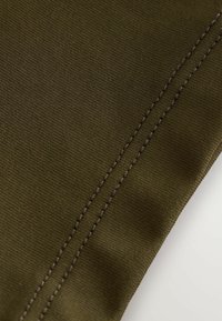 Tmavozelený textil s textúrovaným povrchom, ktorý má výrazný šev prešitý kontrastnou hnedou niťou.