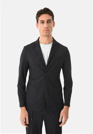 Uomo che indossa un blazer nero aderente sopra una camicia bianca, in piedi con le braccia rilassate contro uno sfondo chiaro e semplice.