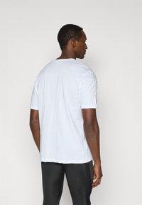 Λευκό μπλουζάκι με κοντά μανίκια, στρογγυλή λαιμόκοψη και υφασμάτινες λεπτομέρειες στους ώμους. Φοριέται με μαύρο αθλητικό παντελόνι.