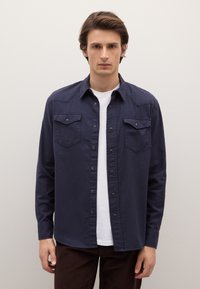 Camisa azul marino abotonada con dos bolsillos en el pecho, cierres a presión y cuello, combinada sobre una camiseta blanca, junto con pantalones marrones.