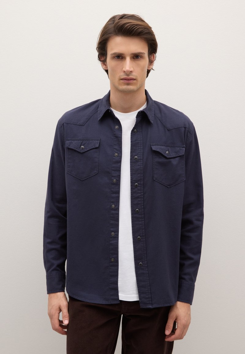 Camisa azul marino abotonada con dos bolsillos en el pecho, cierres a presión y cuello, combinada sobre una camiseta blanca, junto con pantalones marrones.