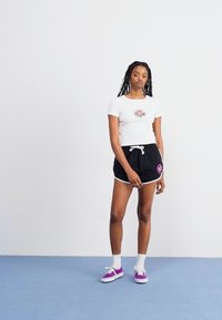 White short-sleeve t-shirt with a floral design, paired with black shorts and purple sneakers, standing on a blue floor against a white wall.