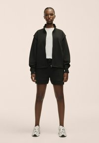 Sudadera negra con cremallera y cuello alto, combinada con pantalones cortos negros. El modelo lleva una camiseta blanca y zapatillas deportivas grises con acentos negros.