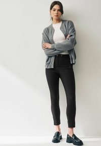 Šedý pletený svetřík, bílý texturovaný top, černé úzké kalhoty a lesklé černé mokasíny. Modelka stojí proti neutrálnímu pozadí.