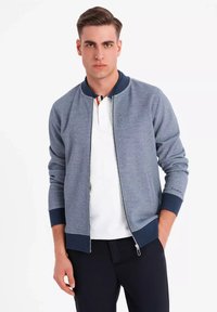 Chaqueta bomber gris con acentos en navy, cierre de cremallera, puños y cinturilla acanalados, confeccionada con una ligera mezcla de algodón. Lleva puesta una polo blanca.
