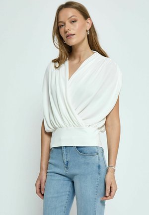Top bianco avvolgente con maniche plissettate e vita aderente, abbinato a jeans azzurri chiari. Tessuto liscio e linee pulite.
