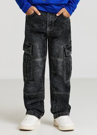 Pantaloni cargo neri con una texture sbiadita, molteplici tasche laterali e cuciture a contrasto, abbinati a sneakers bianche e una maglietta a maniche lunghe blu.