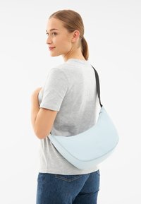 Borsa a spalla azzurro chiaro realizzata in materiale liscio, con design curvo e una tracolla nera, indossata sulla spalla sopra una camicia grigia.