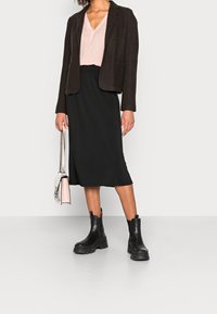 Falda negra con una abertura lateral, blusa de pico rosa claro, chaqueta entallada marrón y botines negros gruesos, junto a un pequeño bolso rosa.