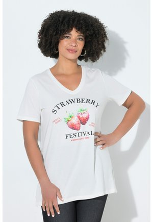 Kvinde med krøllet hår iført en hvid V-hals T-shirt med to jordbær og teksten "Strawberry Festival", stående foran en ensfarvet baggrund.