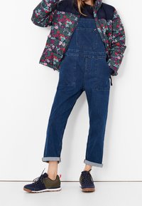 Blomsterprydd pufferjacka i mörkgrön och vinröd, parat med blå denimoverall med två fickor, och marinblå och gröna snörskor.