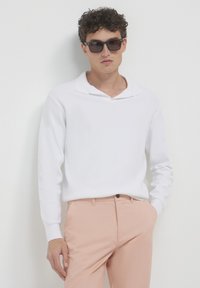 Weißes Langarm-Polohemd mit einem dezenten Kragen, kombiniert mit hellrosa Hosen. Das Modell trägt eine dunkle Sonnenbrille und präsentiert einen sauberen, minimalistischen Look.