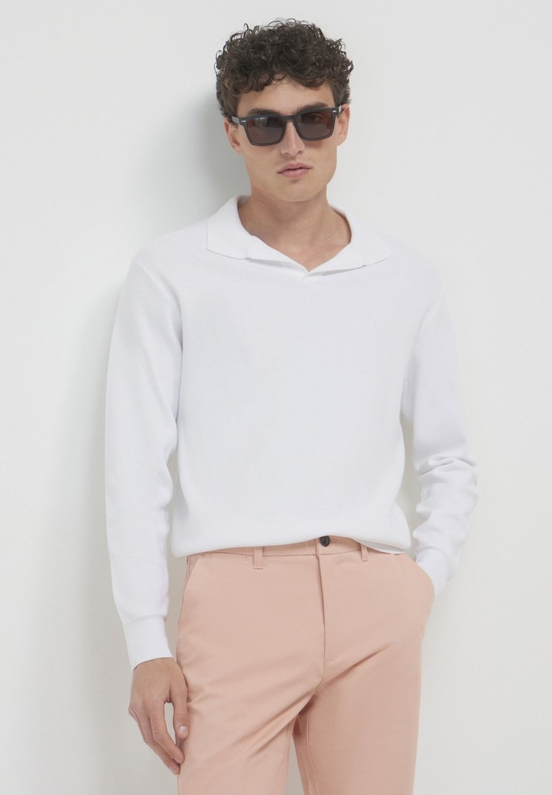Weißes Langarm-Polohemd mit einem dezenten Kragen, kombiniert mit hellrosa Hosen. Das Modell trägt eine dunkle Sonnenbrille und präsentiert einen sauberen, minimalistischen Look.
