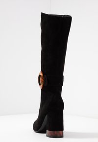 Bota de ante negra hasta la rodilla con punta redondeada, un tacón transparente grueso y un acento circular de color ámbar cerca de la parte superior.