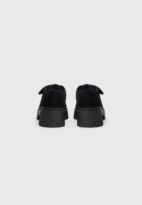Chaussures en daim noir avec une semelle en caoutchouc épaisse. Présentent une forme arrondie et des lacets en tissu. Vue arrière montrant des détails texturés et des coutures.