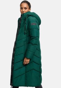 Lunga giacca puffa verde con cappuccio, caratterizzata da un motivo a chevron, una cerniera e bottoni a pressione. Il design include un'etichetta con il logo sulla manica.
