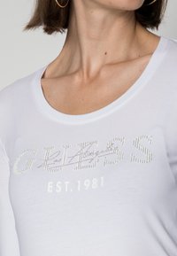 Biała długa koszula z długim rękawem z tekstem z rhinestone "GUESS Los Angeles EST. 1981" oraz okrągłym dekoltem, o gładkiej, lekkiej fakturze materiału.