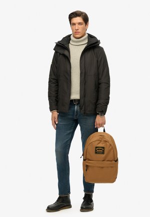 Brun rygsæk med logo, polstret design og frontlomme. Model i sort jakke og lys sweater, blå jeans og sorte sko.