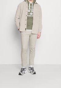 Camicia di corduroy beige sopra una felpa verde con stampa bianca, pantaloni beige chiaro e sneakers nere e argentate. Outfit semplice e casual.