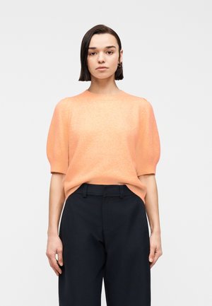 Kvinde med glat mørkt hår iført en ferskenfarvet kortærmet sweater, der er stoppet ned i højtaljede sorte bukser mod en ensfarvet baggrund.