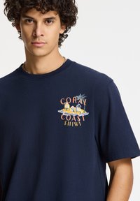 Camiseta de algodón azul marino con un colorido estampado gráfico de casas de playa y palmeras en el pecho. Cuello redondo y mangas cortas.