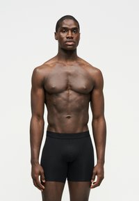 Μυώδης άντρας, όρθιος με το πρόσωπο προς τα εμπρός, φοράει μαύρα ανδρικά εσώρουχα boxer, σε απλό φωτεινό φόντο.