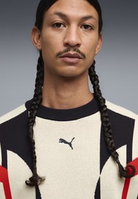 Uomo con due lunghe trecce che indossa un maglione Puma nero, bianco e rosso, rivolto in avanti contro uno sfondo grigio neutro.