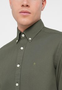 Camisa de color verde oliva con botones blancos, cuello puntiagudo y un pequeño emblema verde en el lado izquierdo del pecho. Textura suave.