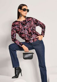 Langärmlige schwarze Bluse mit rotem und lila Paisley-Muster, dunkelblaue Jeans, spitz zulaufende schwarze Stiefeletten und eine kleine schwarze Handtasche mit goldenen Akzenten.