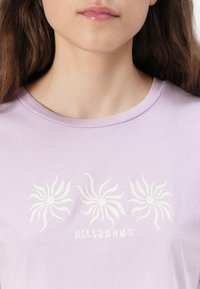 Heledane lilla T-särk pehmest kangast, millel on kolm valget spiraali mustrit ja allpool tekst "BILLABONG" stiliseeritud fondis.