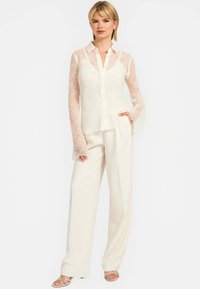 Bluse i blondestof med gennemsigtige lange ærmer og knaplukning fortil, parret med elfenbenfarvede højtsiddende bukser og strop sandaler. Blød, delikat tekstur.