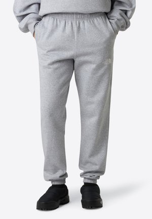 Person trägt graue Sweatpants mit elastischem Bund und Bündchen an den Knöcheln sowie schwarze Slipper, die Hände in den Taschen, steht vor weißem Hintergrund.