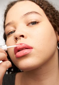 Lipglossapplicator wordt gebruikt op volle lippen. De gloss heeft een lichtroze kleur met een glanzende afwerking. De productverpakking is zichtbaar nabij de hand.